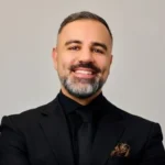 Man with beard wearing a black suit and tie with a patterned pocket square, smiling.