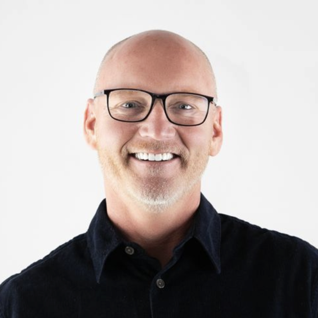 Bald man with glasses and beard smiles wearing a dark shirt