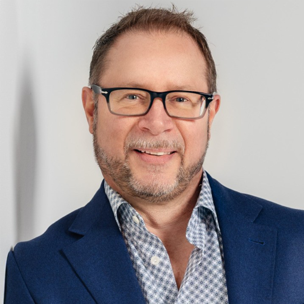 Man with glasses and goatee wearing a blue blazer and patterned shirt smiles at camera