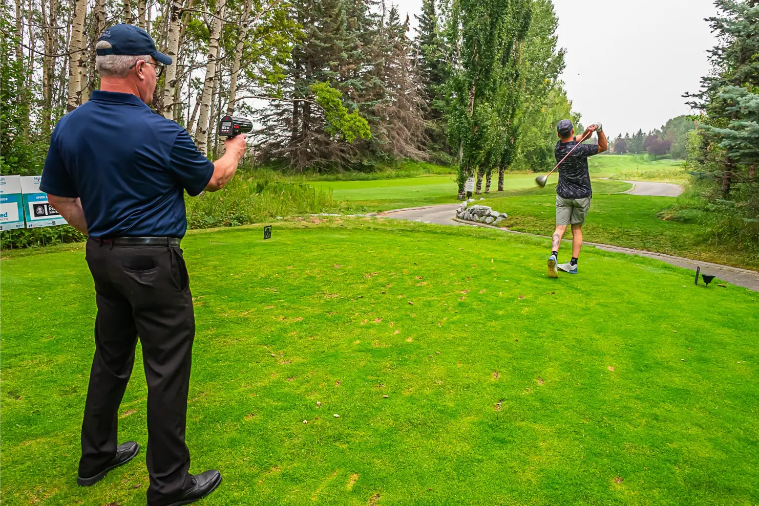 Golfer swings driver as a man with a laser rangefinder watches on a grassy tee box.