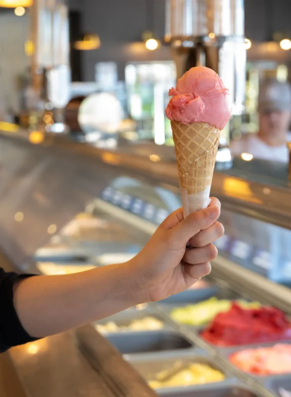 Hand holding pink strawberry ice cream cone with gelato display case in background