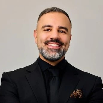Man in a black suit and tie with a grey beard smiles.