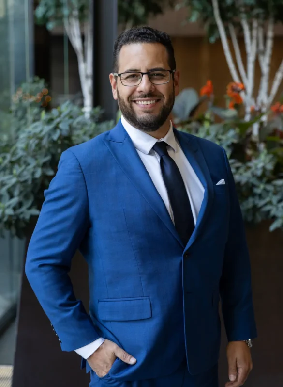Man in blue suit with tie and glasses smiles, hand in pocket.