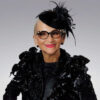 Fashionable older woman in black feathered hat and ornate glasses.
